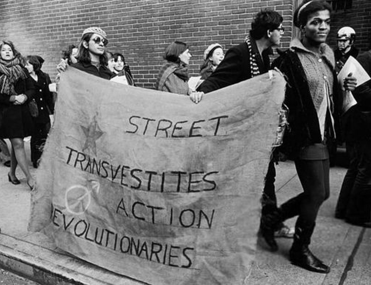 Imagem de uma manifestação com pessoas vestindo roupas características dos anos 60 e 70. As vestimentas incluem saias curtas, botas, casacos de lã e acessórios como óculos escuros e chapéus. Dia do Orgulho LGBTQIA+.