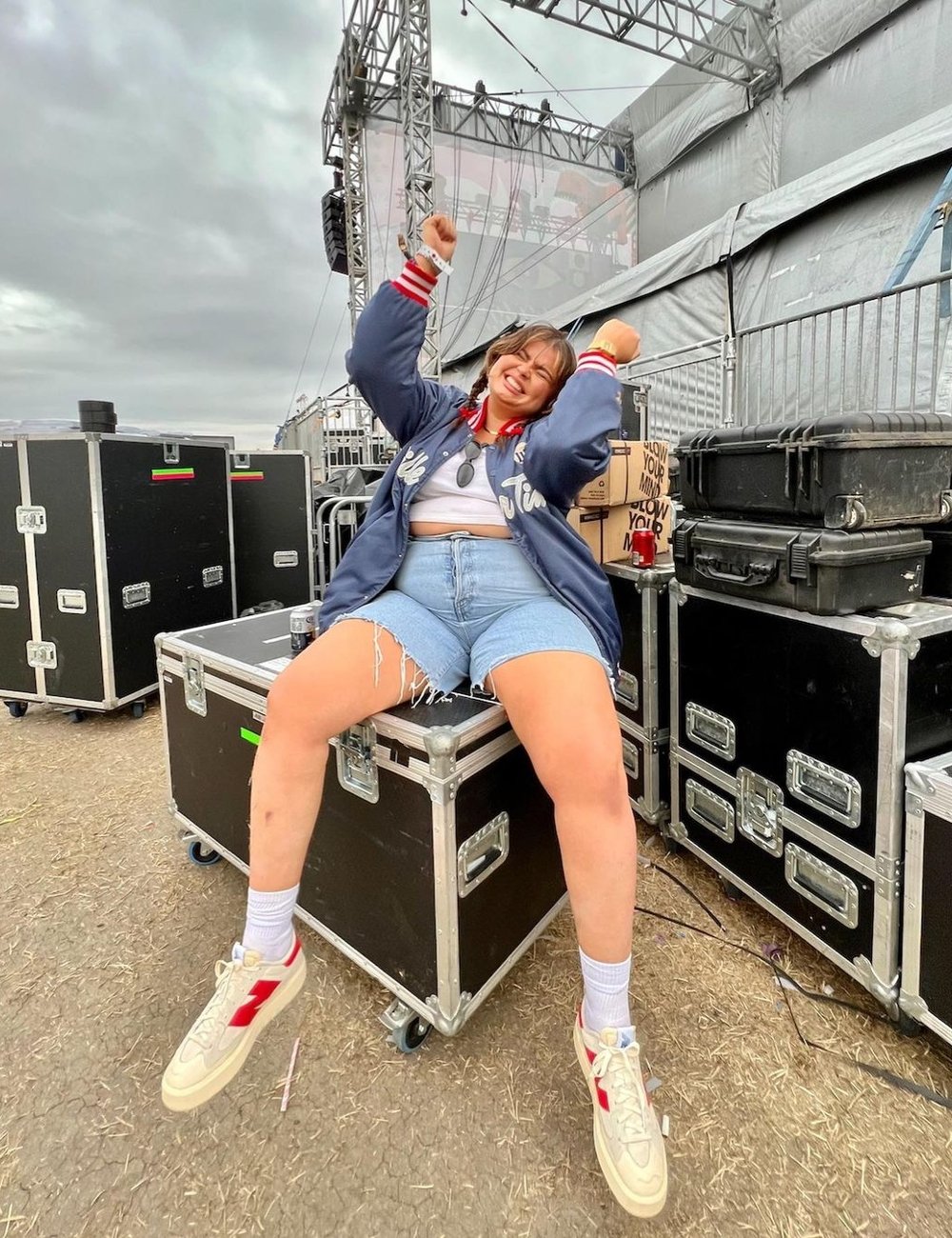 Pessoa sentada em caixa de equipamento no backstage de um show, vestindo jaqueta oversized estilo varsity, cropped branco, bermuda jeans desfiada e tênis brancos com detalhes vermelhos. Look casual e despojado, popular em um festival de música.