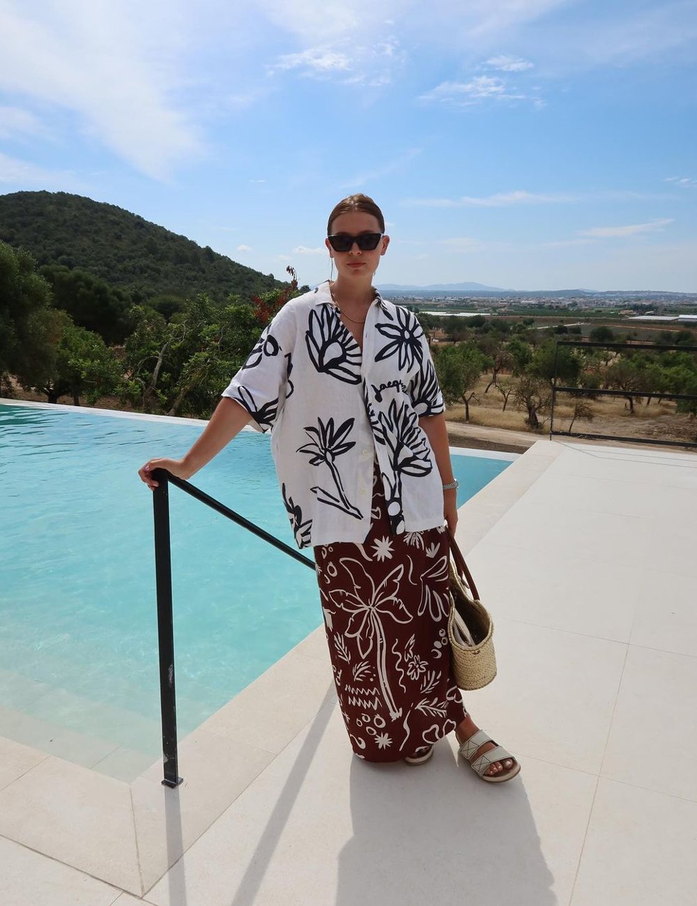 Pessoa em look casual junto a uma piscina, usando camisa estampada com flores grandes em preto e branco e saia longa marrom com design floral. Complementa o visual com sandálias brancas, bolsa de palha e óculos de sol. Fim de semana vai ter sol.