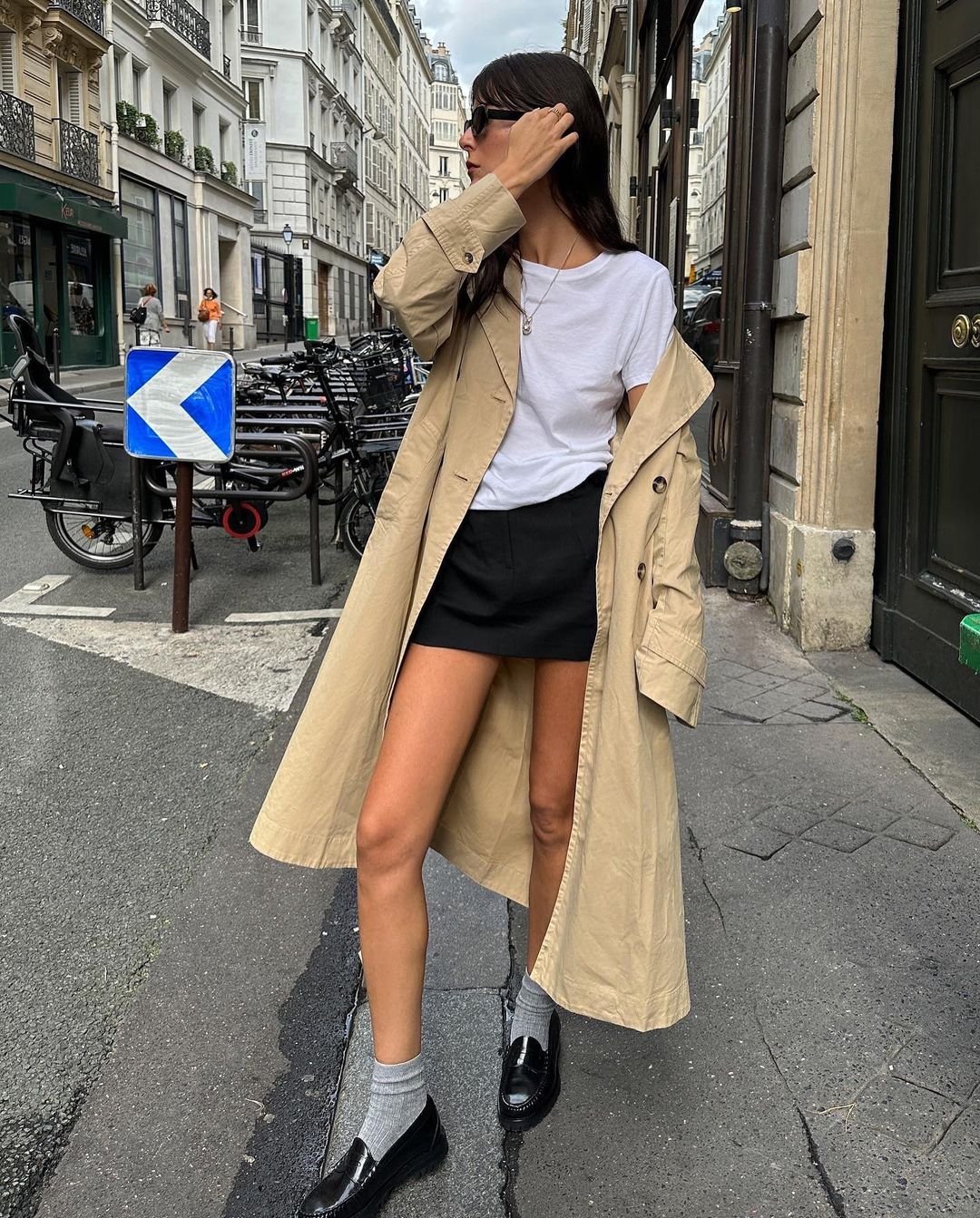 Rua urbana, mulher vestindo trench coat bege, camiseta branca básica, saia preta curta, mocassim preto e meia cinza. Tendência de moda minimalista e elegante, ideal para meia-estação. Kombinação de cores neutras e estilo casual-chic.