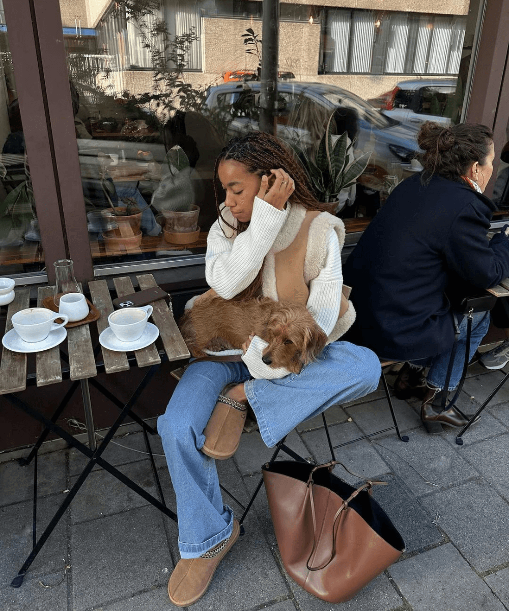 Amaka Hamelijnck - calça jeans, blusa trico off white, colete e botas ugg - looks para festa junina com calça jeans - inverno - mulher negra de tranças sentada na rua segurando um cachorro caramelo no colo - https://stealthelook.com.br