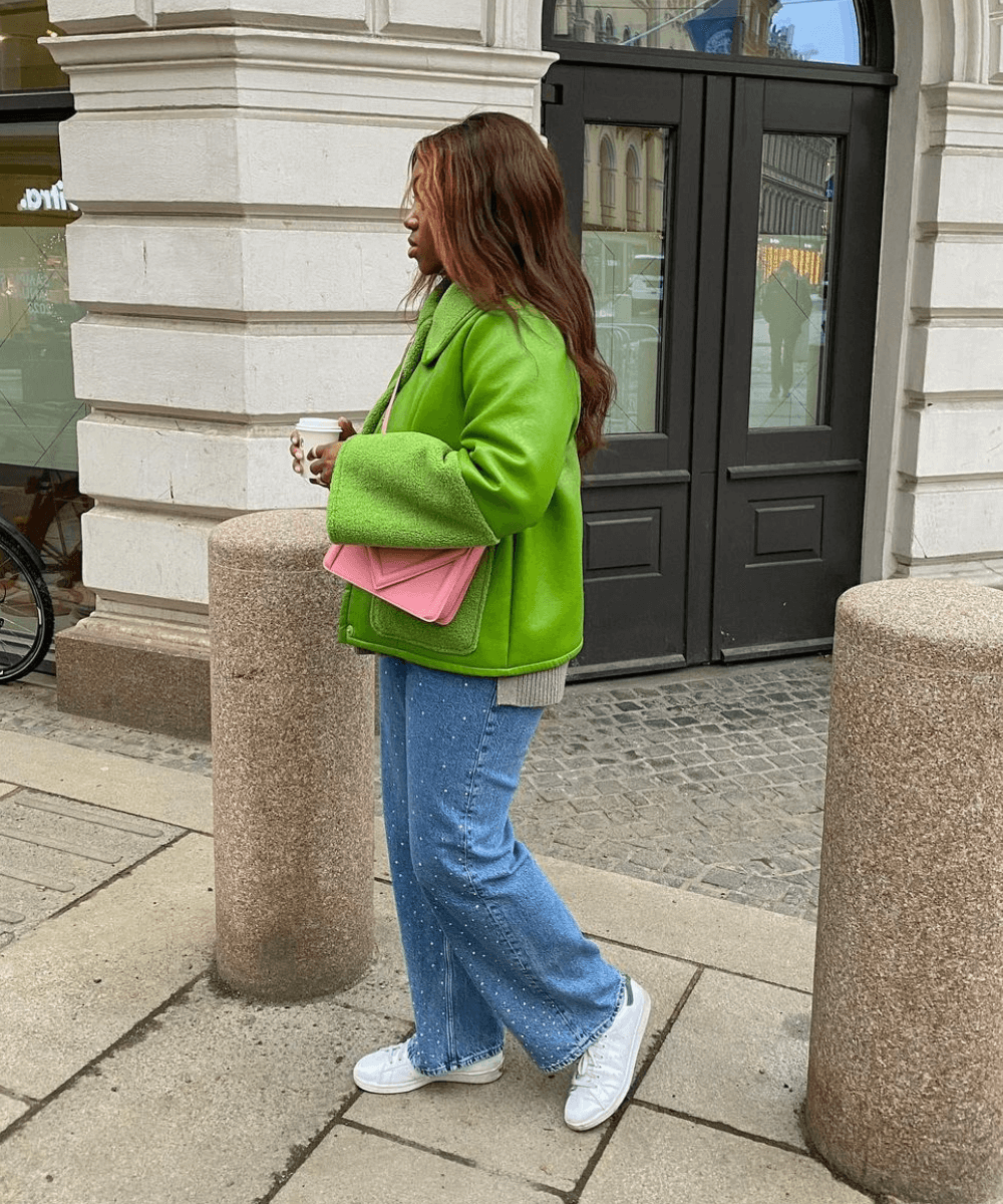 Nnenna Echem - calça jeans, jaqueta verde, bolsa rosa e tênis - looks para festa junina com calça jeans - inverno - mulher negra em pé na rua de lado - https://stealthelook.com.br