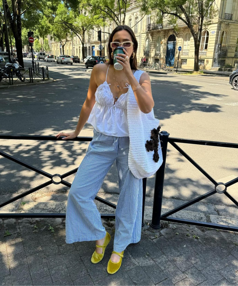 Mulher posando na rua usando blusa branca de alças finas e babados, calça pantalona listrada azul e branca, sapatos verdes de veludo e bolsa de crochê branca. Óculos escuros e acessórios dourados compõem o look casual e moderno.