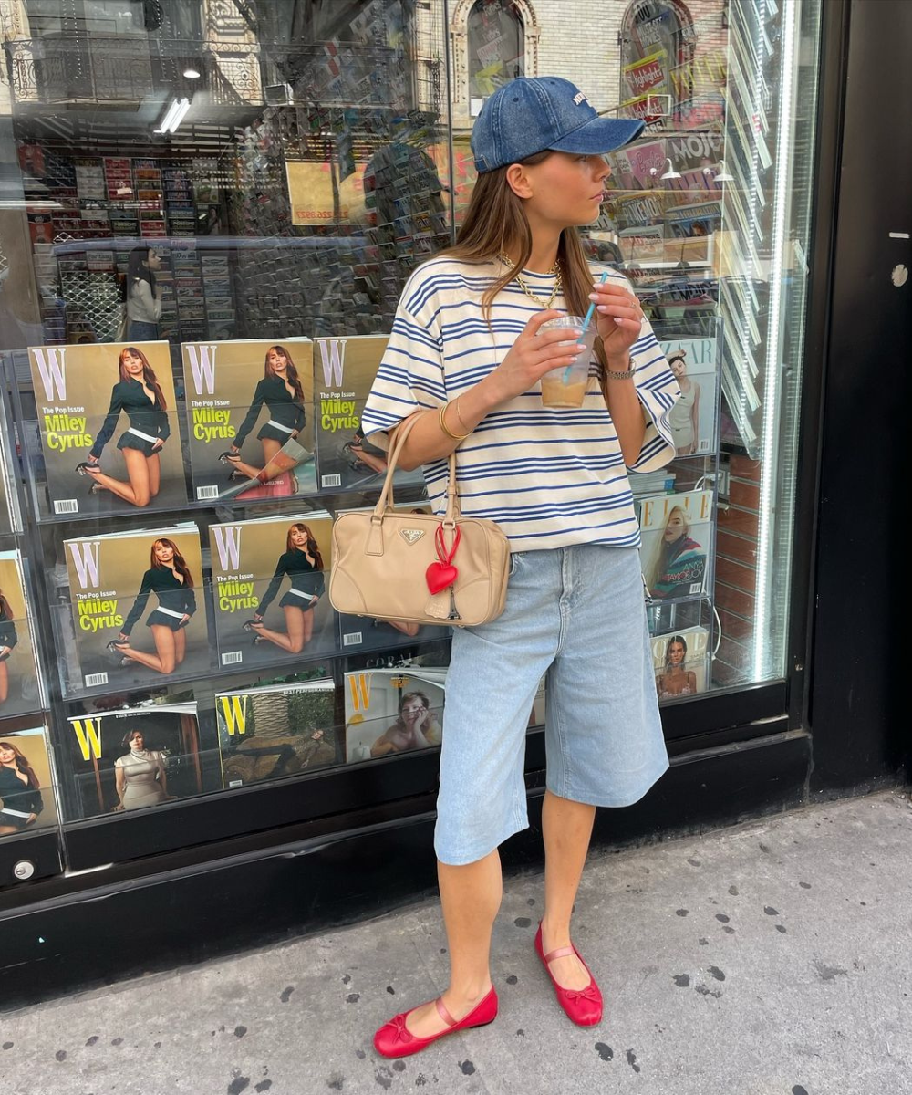 Mulher vestindo camisa listrada, bermuda jeans, sapatilhas vermelhas e boné azul. Acessórios incluem bolsa bege e colares. Moda casual contemporânea, com influência de estilo náutico e elementos coloridos.