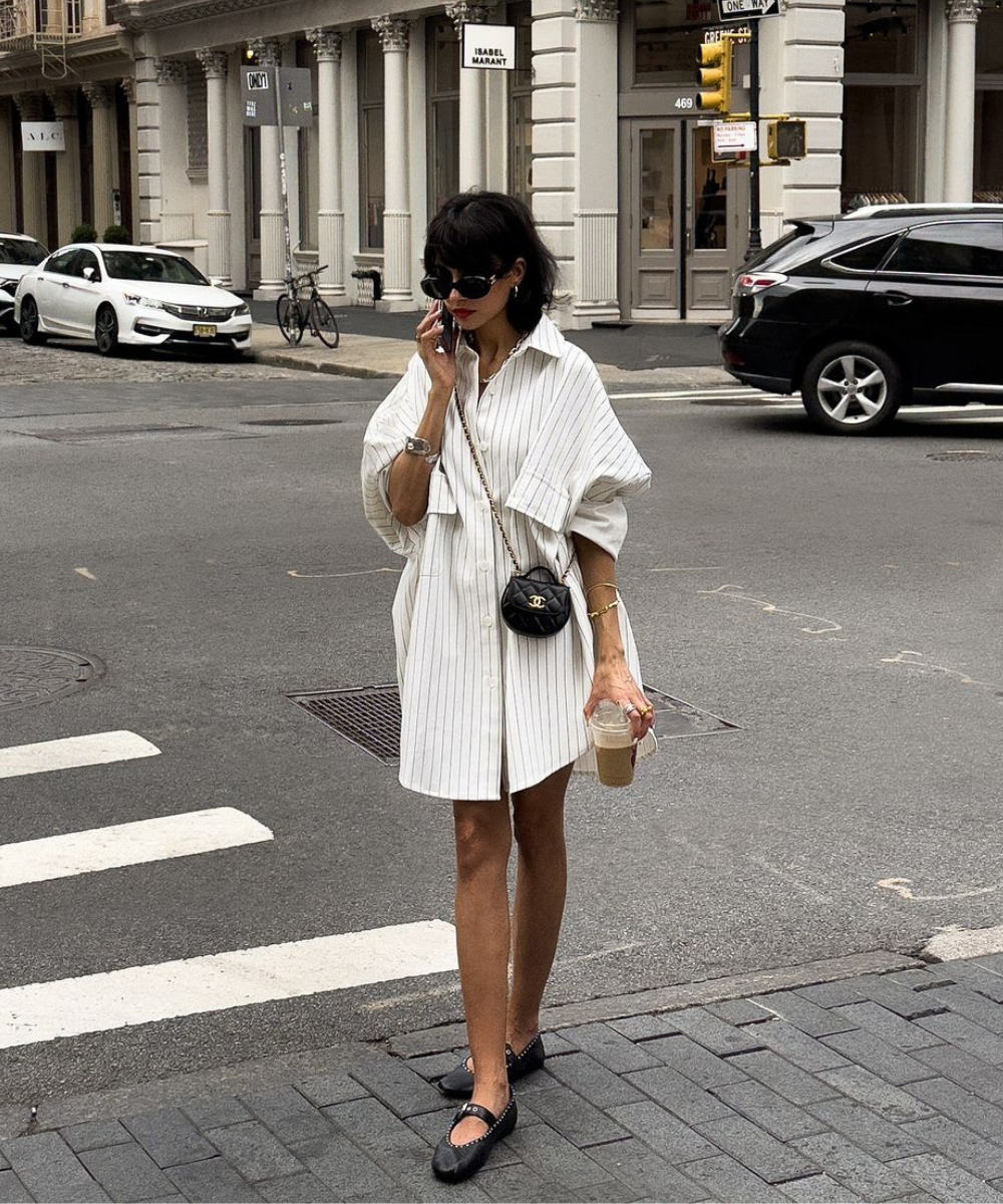 Mulher com vestido-camisa listrado branco oversize, mini bolsa preta a tiracolo, óculos de sol grandes e sapatilhas pretas. Visual casual e sofisticado, com acessórios minimalistas, refletindo tendências de moda urbana contemporânea.