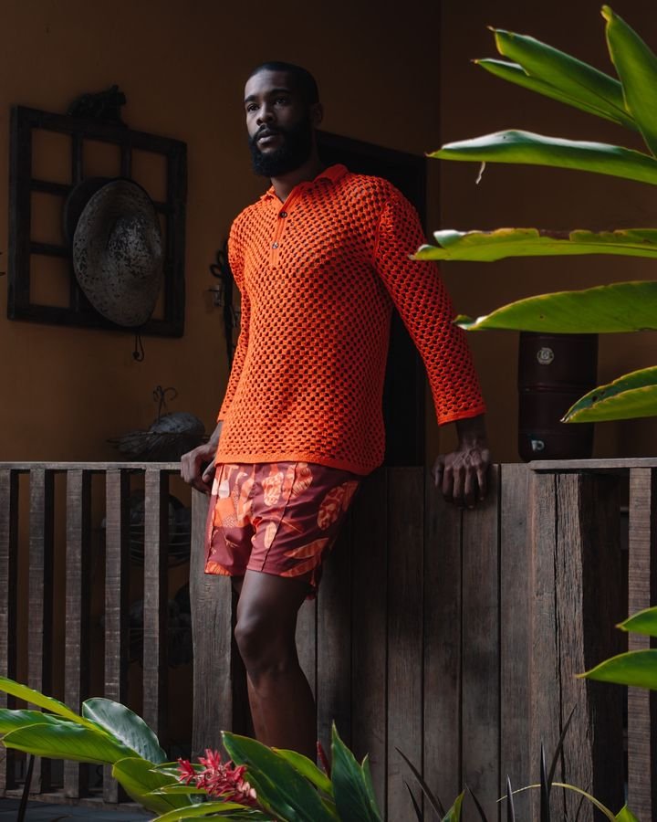 Homem com um estilo vibrante e contemporâneo. Veste uma camisa laranja vazada de manga longa, combinada com um short estampado em tons de laranja e marrom. Tendência que destaca texturas e cores ousadas, ideal para climas tropicais.