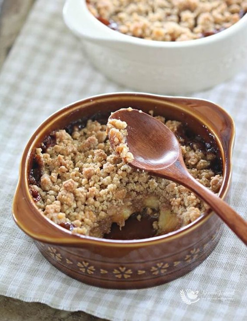 Sobremesa crumble de banana com óleo de coco. Teve uma mordida retirada, revelando o recheio de frutas sob a cobertura crocante. Fundo de tecido de linho bege com listras. Ideal para indexação de receitas e pratos caseiros.