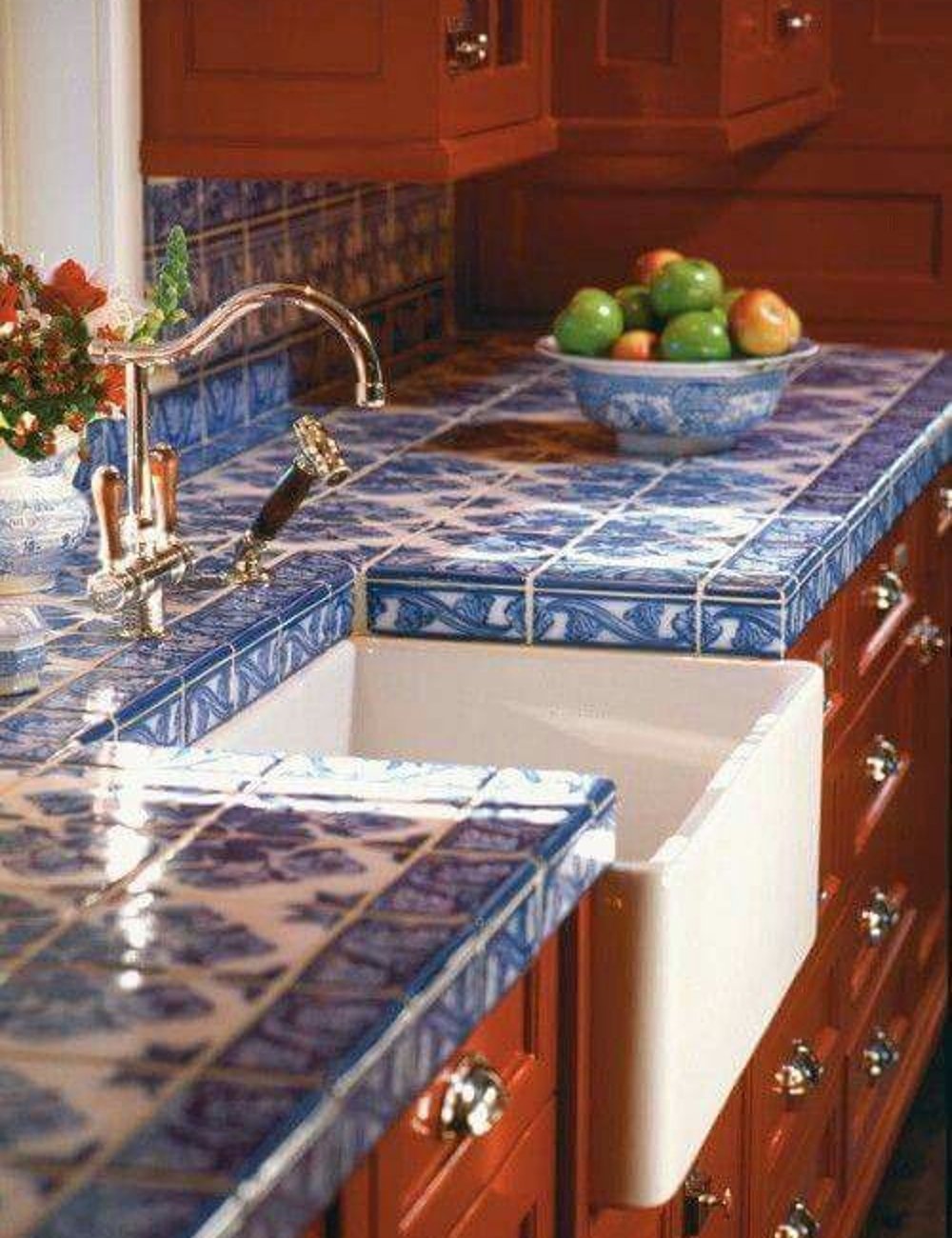 Pias de cozinha com decoração rústica e sofisticada, destacando azulejos azuis com padrões florais e armários de madeira vermelha. Pia estilo fazenda branca e torneira clássica. Fruteira de cerâmica azul com maçãs e laranjas no balcão.