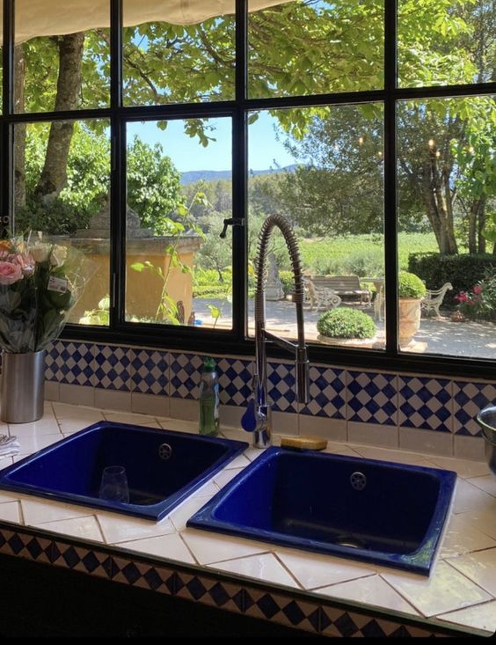 Pias de cozinha dupla azul, bancada de ladrilhos brancos e azulejos decorativos em tons de azul e branco. Janela ampla com vista para jardim verdejante e montanhas ao fundo.