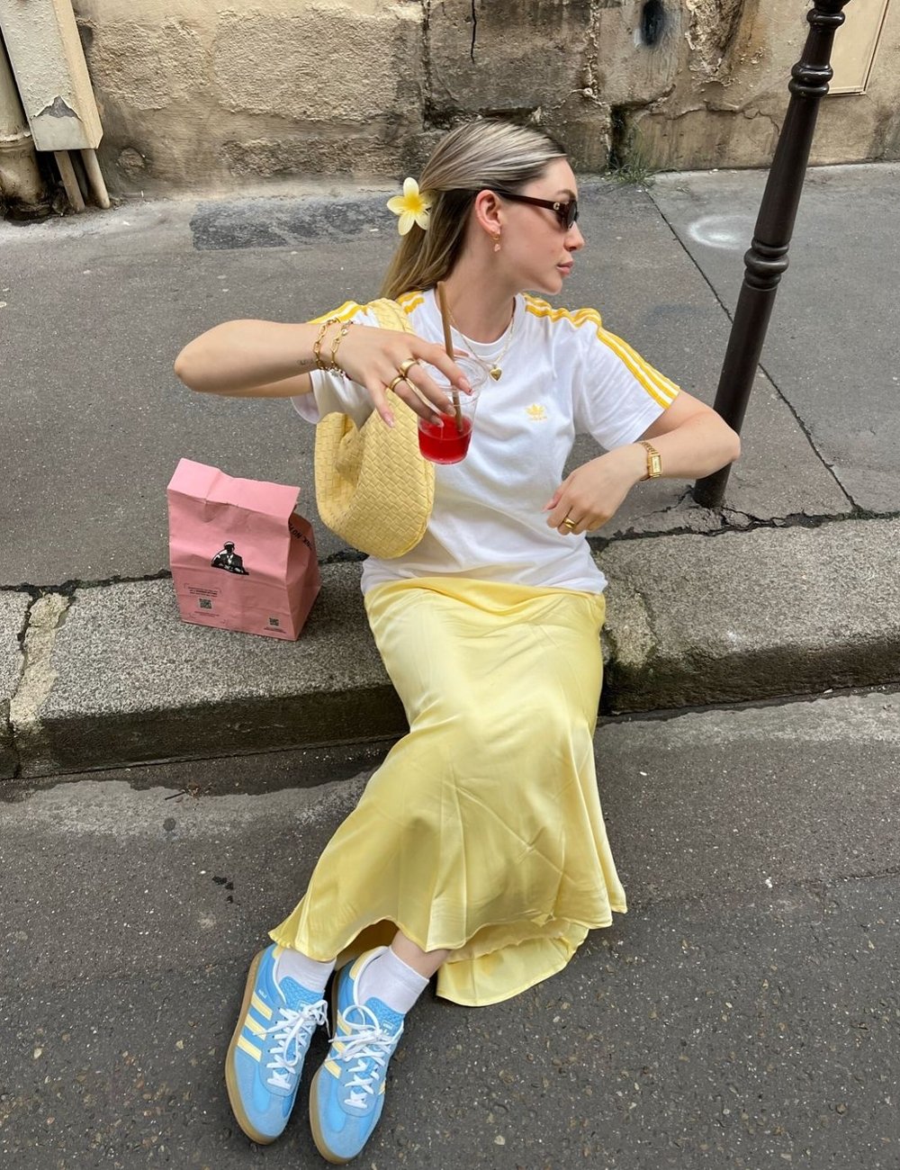 Mulher sentada, vestindo uma camiseta branca com detalhes amarelos, saia midi amarelo claro e tênis azuis com listras amarelas. Acessórios: bolsa de palha, óculos de sol, flores no cabelo e joias douradas. Tendências de moda de 2024.