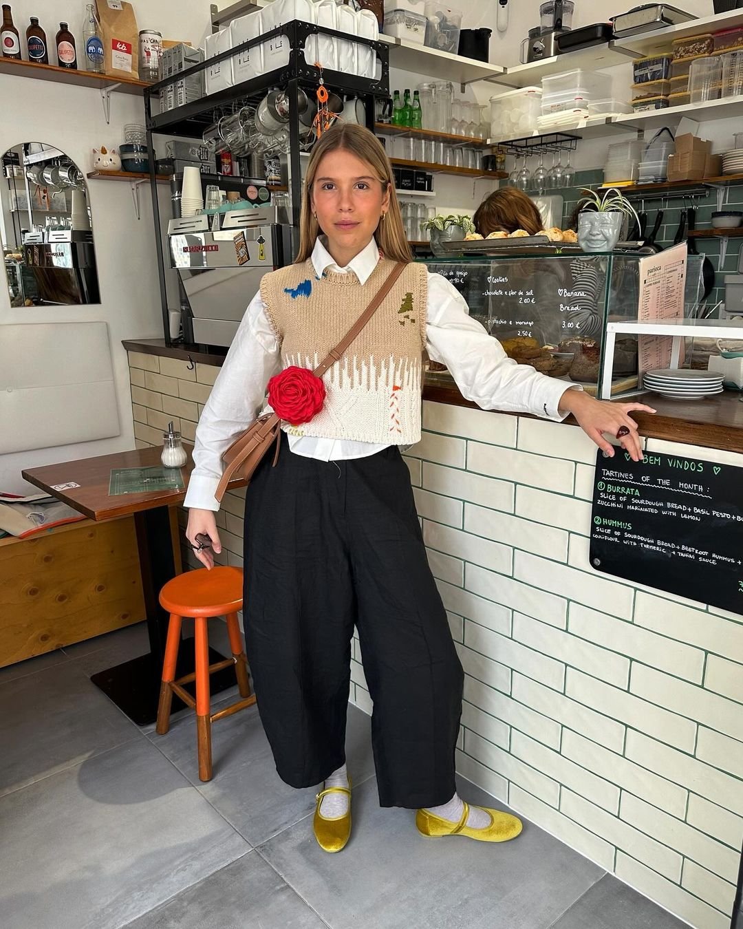 Jovem usando um colete bordado sobre camisa branca, acompanhada de calça preta solta e sapatilhas amarelas vibrantes. O look é complementado por acessórios, incluindo uma bolsa pequena e uma flor vermelha como detalhe decorativo. atmosfera casual-chic.