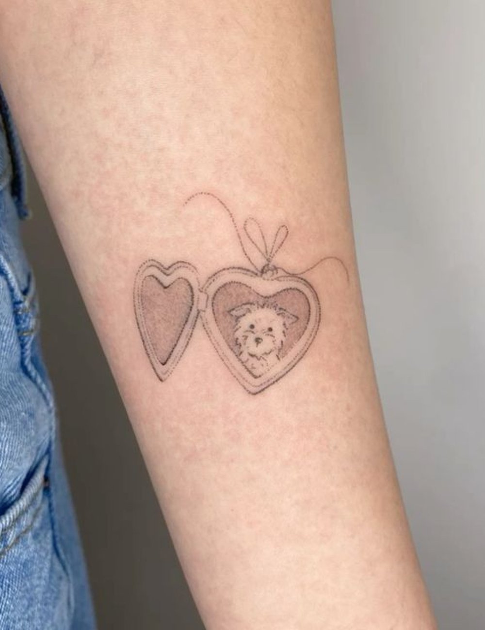 Tatuagem delicada no braço de um medalhão em forma de coração com retrato de cachorro. A pessoa veste jeans claros, seguindo a tendência casual e atemporal. Moda minimalista e personalizada.