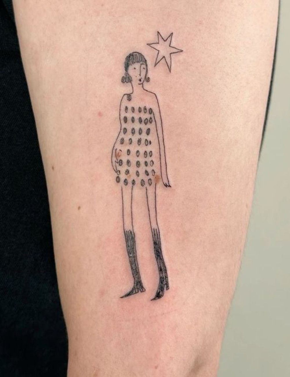 Tatuagem minimalista de figura estilizada com vestido curto de bolinhas e botas longas, ao lado de uma estrela. O desenho combina simplicidade e elegância, refletindo tendências modernas de moda e arte corporal.