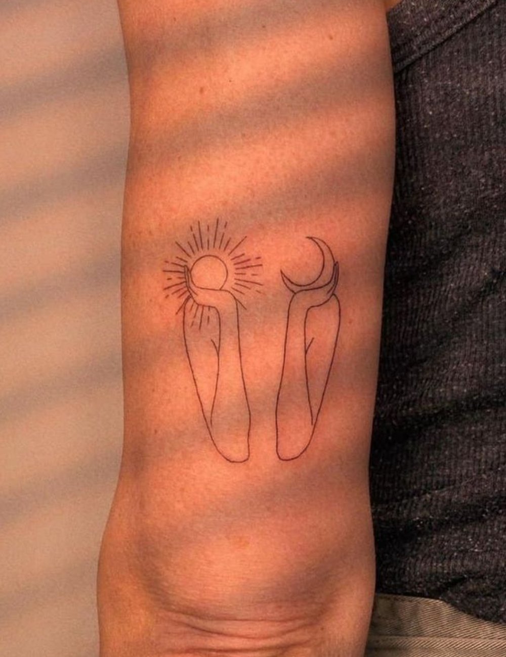 Tatuagem de linha fina com desenhos minimalistas de mão segurando sol e lua no braço. Tendência em alta entre os jovens adeptos de arte corporal delicada. Vestimenta casual com camiseta cinza básica.