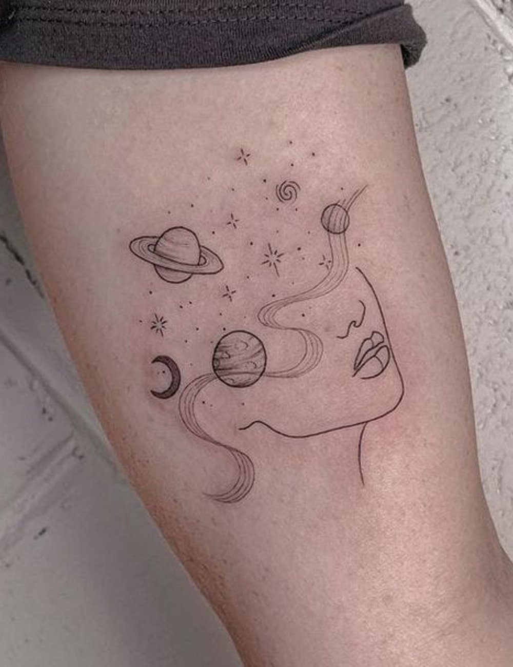 Tatuagem minimalista na coxa com tema espacial e elementos de planetas e estrelas. Design artístico combina rosto feminino com linhas suaves e astros. Tendência de tatuagens de linha fina e temática cósmica.