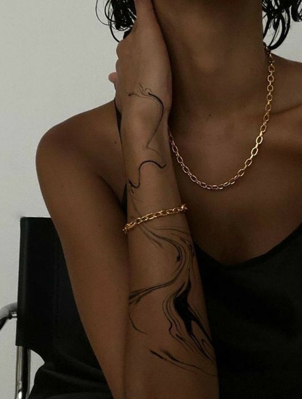 Mulher usando colar e pulseira de corrente dourada. Ela veste uma blusa de alça preta que deixa parte do ombro nu. No braço, um desenho artístico com linhas fluídas que lembra uma tatuagem temporária. Estilo minimalista e elegante.