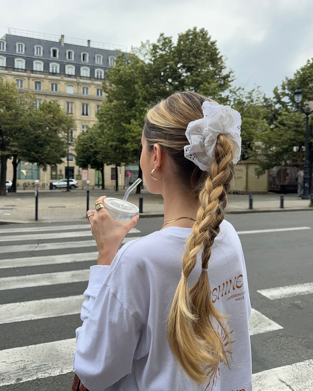 Mulher usando camiseta branca oversized com estampa nas costas, segurando bebida com canudo. cabelo trançado adornado com scrunchie branca, além de acessórios como brincos de argola e anéis dourados. Estilo casual e moderno. acessórios de cabelo