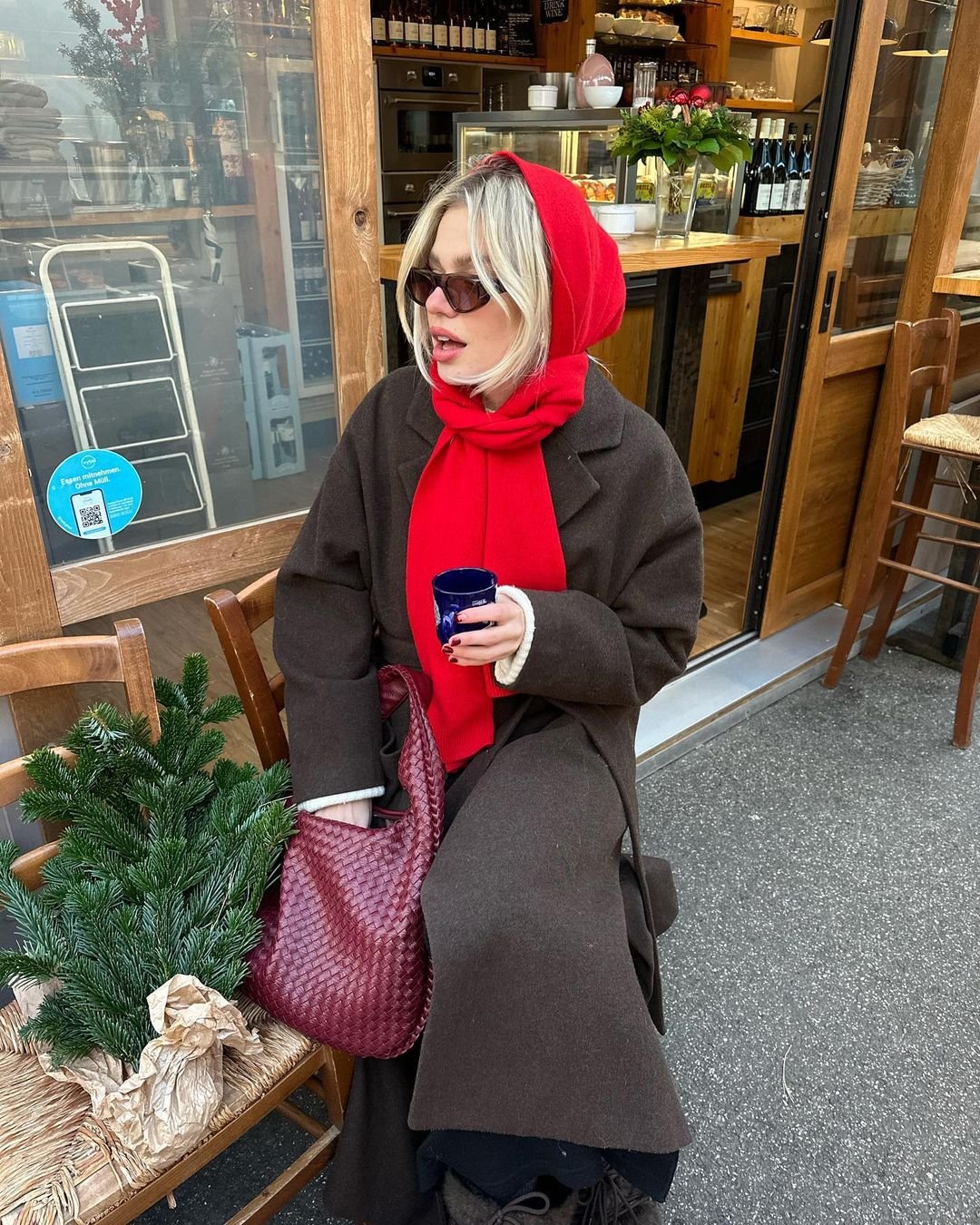 Pessoa sentada em um café vestindo um sobretudo marrom, cachecol vermelho usado como capuz, óculos de sol e carregando uma bolsa vermelha de textura trançada. A moda destaca um estilo de inverno casual chic com foco em acessórios vibrantes.