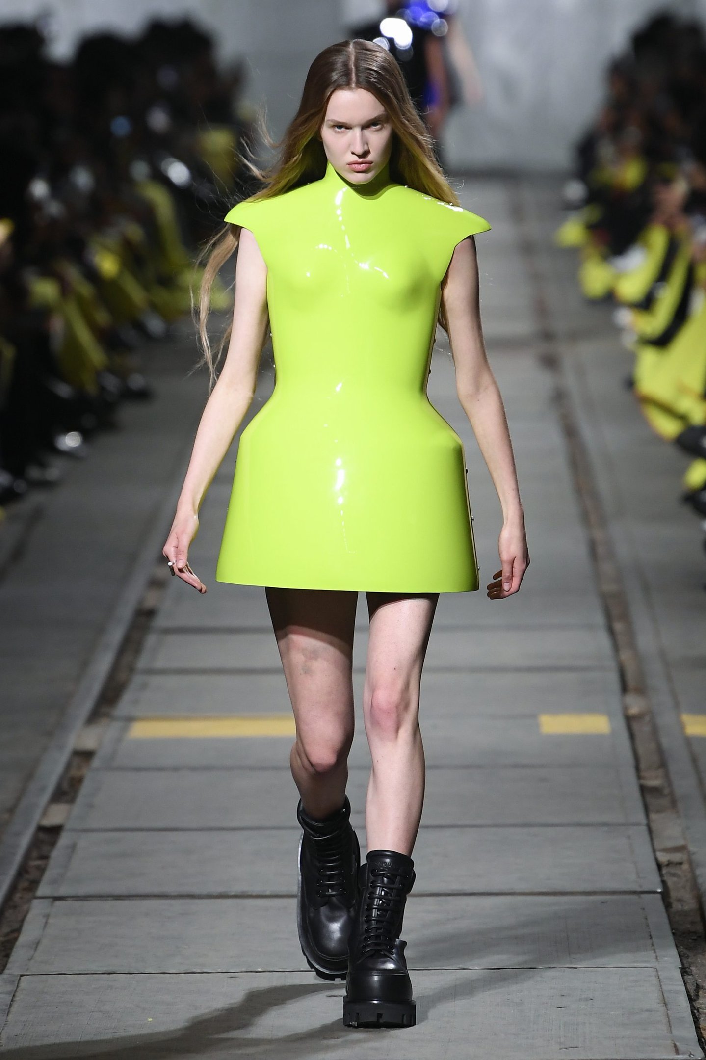 Modelo desfilando em uma passarela, vestindo um mini vestido verde neon de couro estilo futurista e botinhas pretas robustas. Look destacando tendências de moda vanguardista e ousada, combinando cores vibrantes com elementos de design contemporâneos.