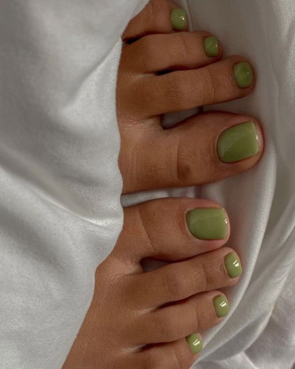 Unhas dos pés pintadas com esmalte verde oliva, sob um fundo de tecido branco. A tendência de esmalte colorido para unhas dos pés está em alta, sendo uma escolha moderna e elegante que adiciona sofisticação e personalidade ao look.