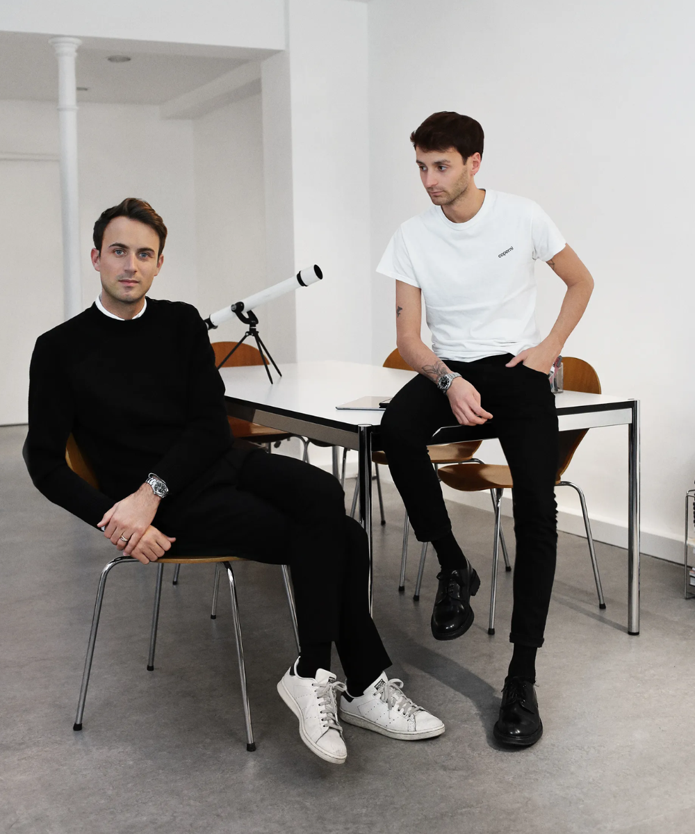 Dois homens em uma sala minimalista. Um veste suéter preto e calça preta com tênis branco, estilo casual sofisticado. O outro usa camiseta branca e calça preta com sapatos pretos, combinando conforto e elegância contemporânea.