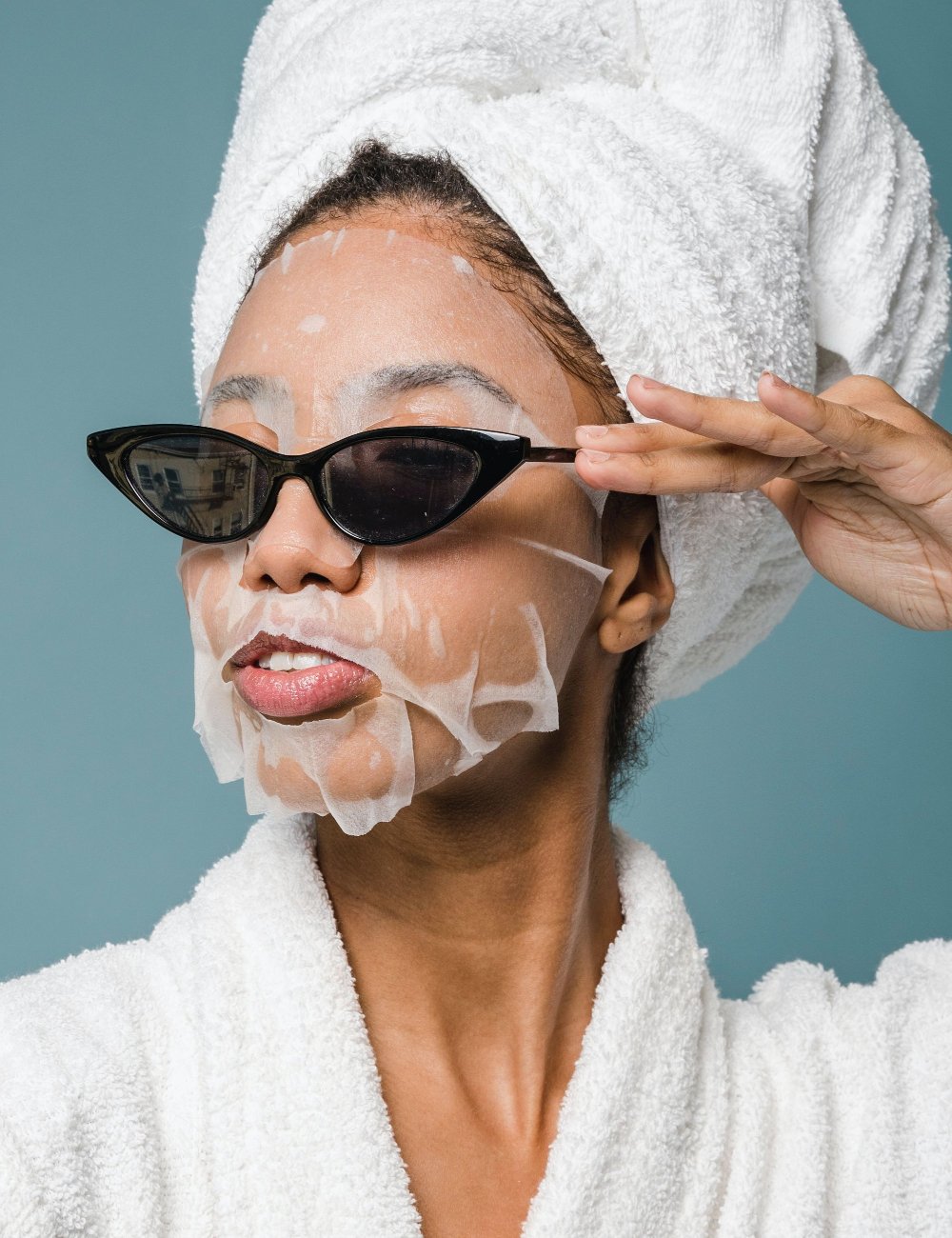 Pessoa vestindo roupão branco e toalha na cabeça, usando máscara facial e óculos de sol estilo retrô. Combinação de autocuidado e moda, tendência popular em cuidados com a pele e estilo casual-chic. Fundo azul claro. Melhorar a autoestima.