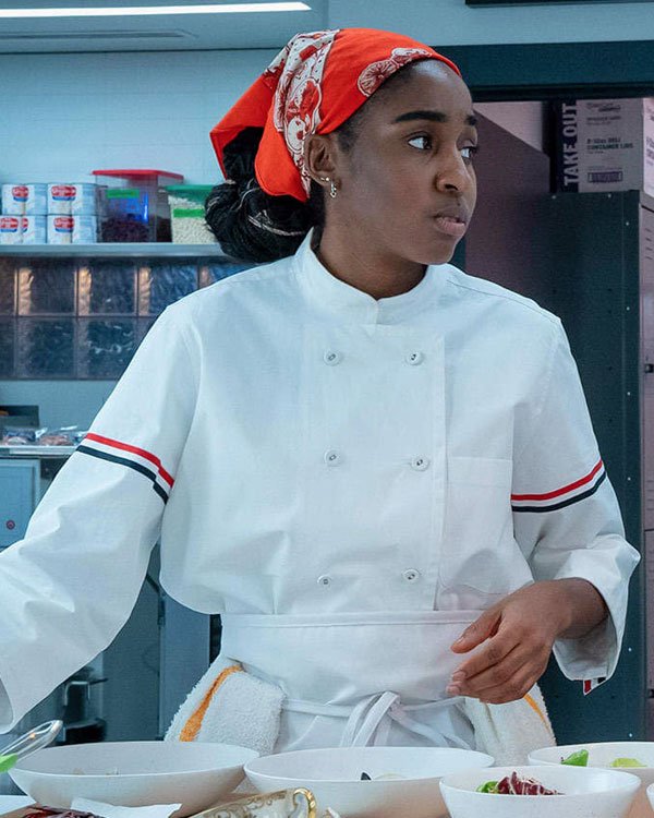 Jovem chef de cozinha vestindo um uniforme branco tradicional de chef, com listras coloridas nos ombros. Ela usa um lenço vermelho decorado na cabeça, adicionando um toque de estilo pessoal. A moda na culinária destaca a praticidade com identidade visual.