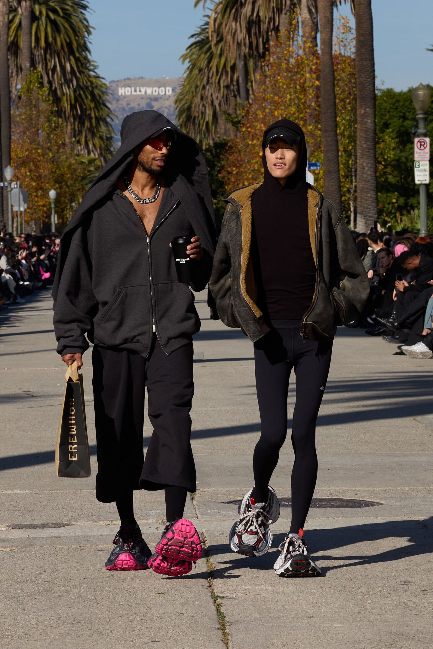 Dois homens caminham vestidos de forma casual e moderna. Um usa moletom com capuz, calça larga e chinelos de pelúcia rosa. O outro veste jaqueta de couro, touca, leggings e tênis esportivos. Ambos exibem acessórios estilosos como óculos escuros e colares.