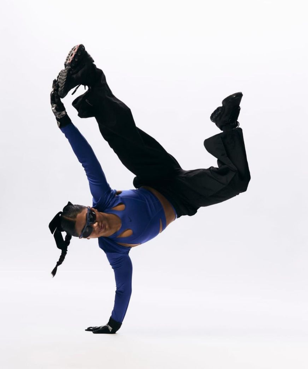 Mulher dançando break dance vestindo uma blusa de manga longa azul com recortes modernos, calça larga preta e luvas esportivas. Acessórios como óculos escuros e cabelo preso com laços, transmitindo um visual dinâmico e contemporâneo.