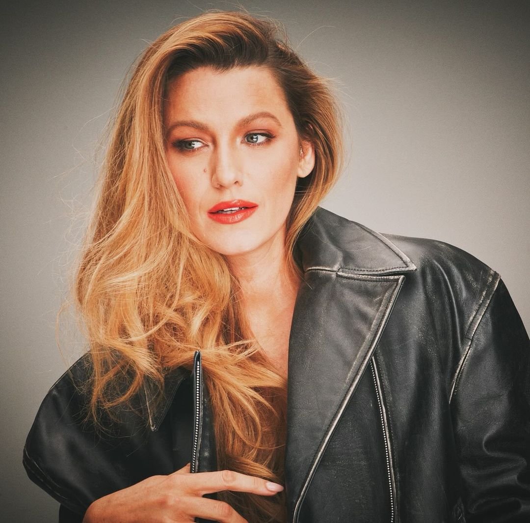 Mulher usando jaqueta de couro preta, com cabelo loiro volumoso e maquiagem destacando batom vermelho e sombra suave. Estilo rock glam, tendência atual em moda que mistura atitude e sofisticação. blake lively