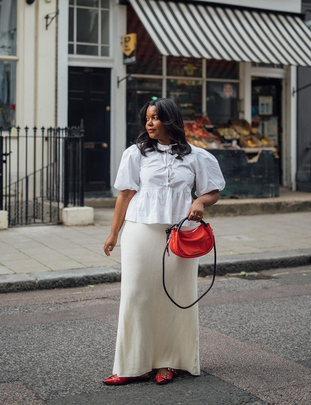 Mulher veste blusa com amarração e babados, combinada com saia longa branca. Completa o look com sapatos e bolsa vermelhos, destacando-se pela paleta de cores contrastantes e tendência oversized.