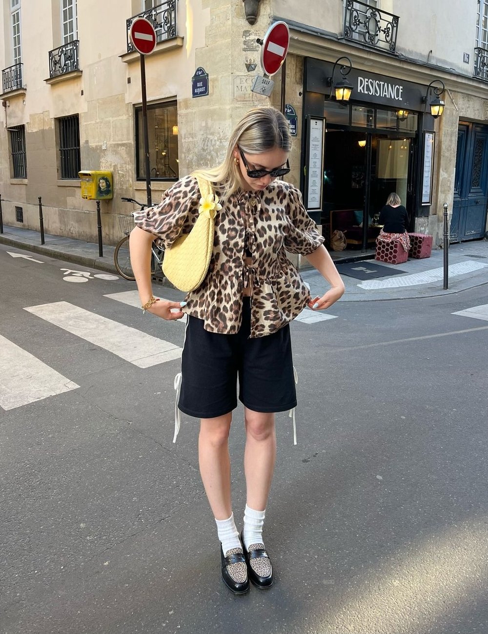 Pessoa vestindo blusa com amarração, shorts preto e sapatos pretos com detalhes de estampa animal. Acessórios incluem bolsa amarela e óculos de sol. Look moderno e casual, combinando conforto com tendências de moda animal print e cores básicas.