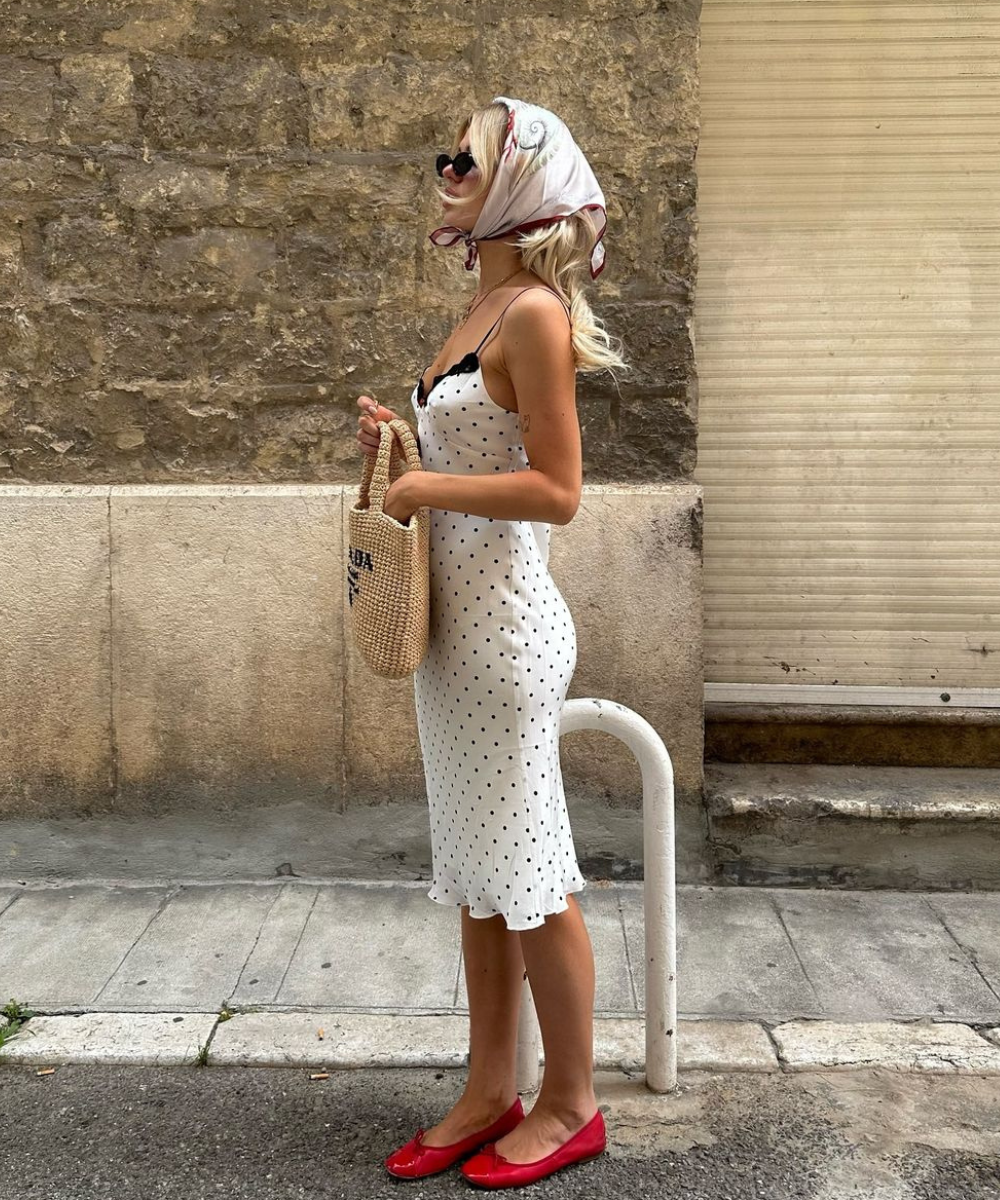 Mulher vestindo vestido midi de alças branco com bolinhas pretas, lenço na cabeça, óculos de sol, bolsa de palha e sapatilhas vermelhas. Look elegante e vintage com fortes referências retrô, ideal para dias de verão.