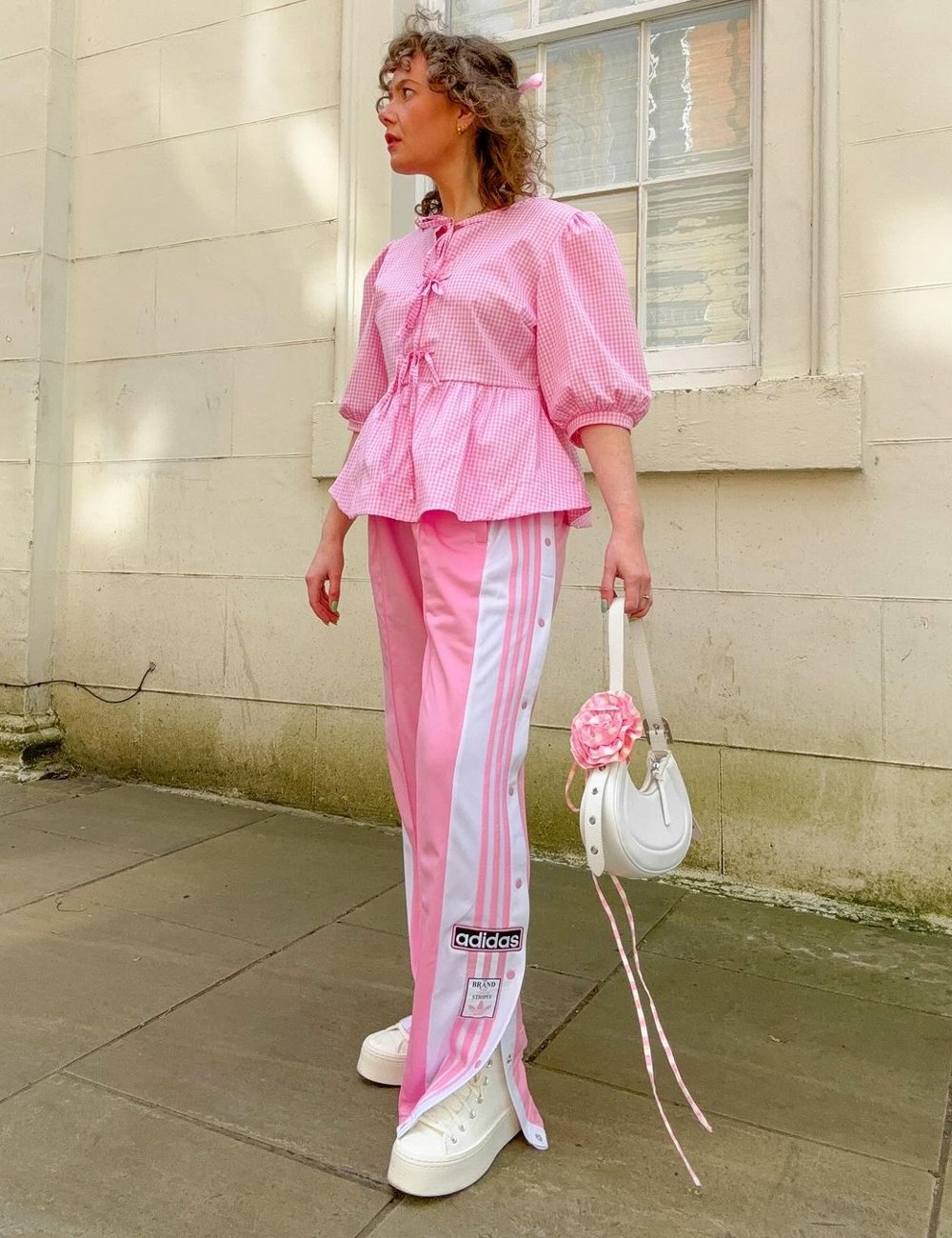 Pessoa vestindo blusa rosa de mangas bufantes e calça esportiva rosa com listras brancas da Adidas; bolsa meia-lua branca decorada com flor rosa e tênis branco. Look combina elementos românticos com moda esportiva.