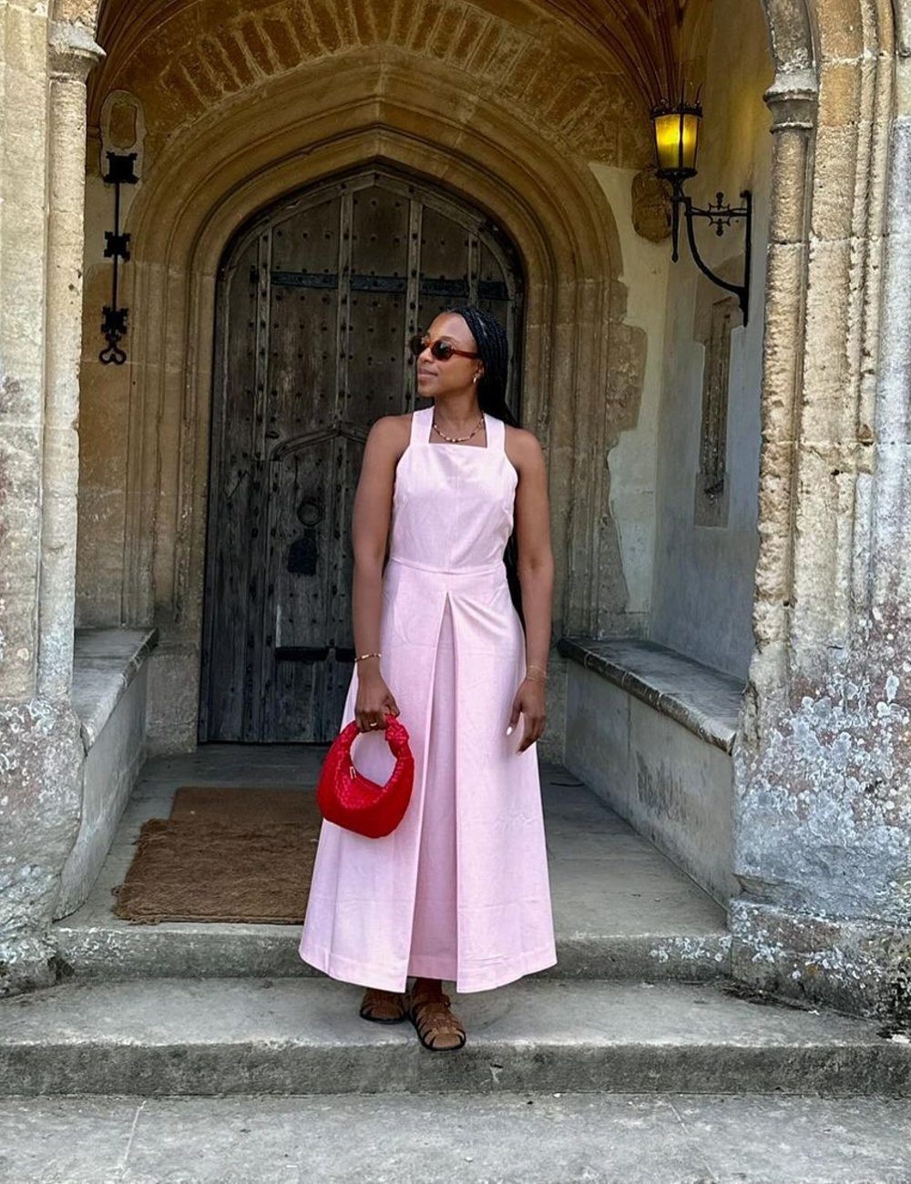 Mulher usando vestido rosa claro midi, com modelagem solta e alcinhas, combinando com bolsa meia-lua vermelha de mão e sandálias rasteiras marrons. Óculos escuros completam o look casual e elegante, ideal para um dia ensolarado.