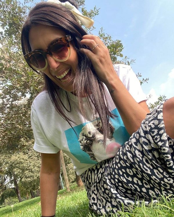 Mulher usa camiseta branca com estampa de arte, óculos de sol oversized, saia de padrão animal print e laço no cabelo, evidenciando um estilo casual e despojado. Tendência de moda inclui mix de estampas e acessórios retro.