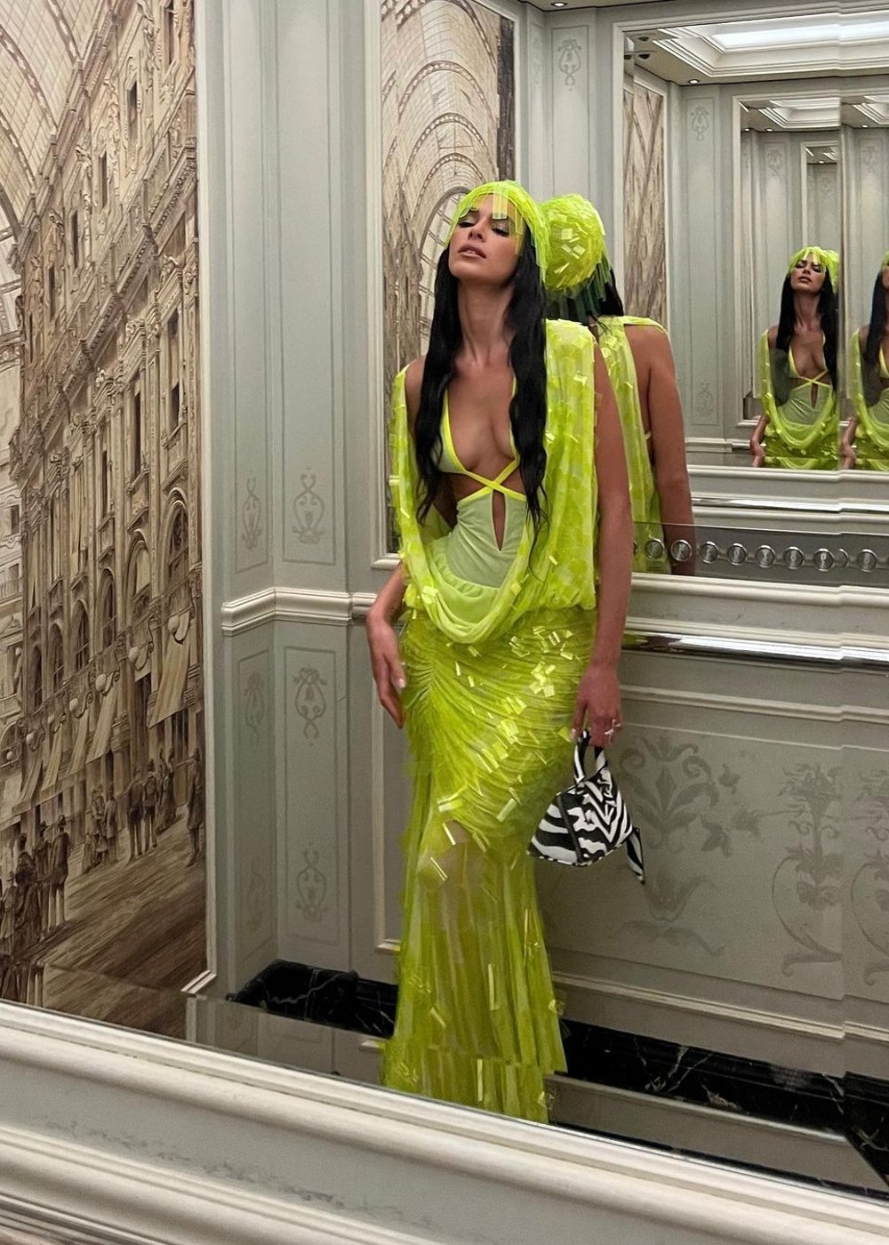 Bruna Marquezine vestindo um conjunto neon verde com transparências e franja, incluindo um turbante da mesma cor. O top tem um design recortado com alças cruzadas. Ela carrega uma bolsa de estampa zebrada.