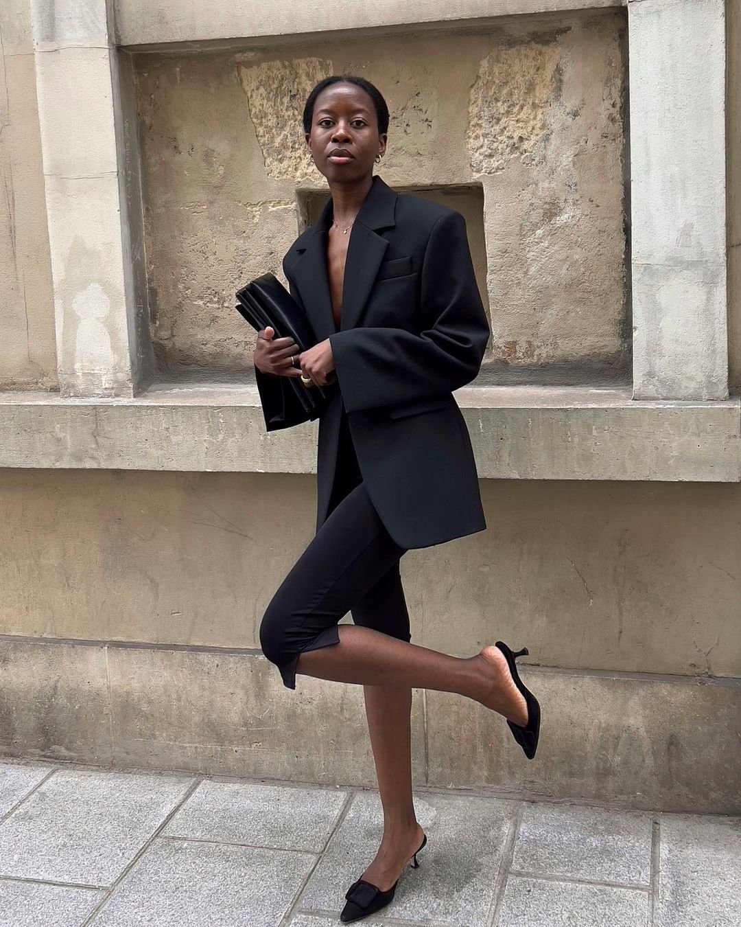 Mulher vestindo blazer oversized preto, calça capri preta e mule de salto baixo. Look elegante e minimalista, combinando tendências de alfaiataria com peças esportivas. Ela segura uma clutch preta, criando um visual chic descomplicado.