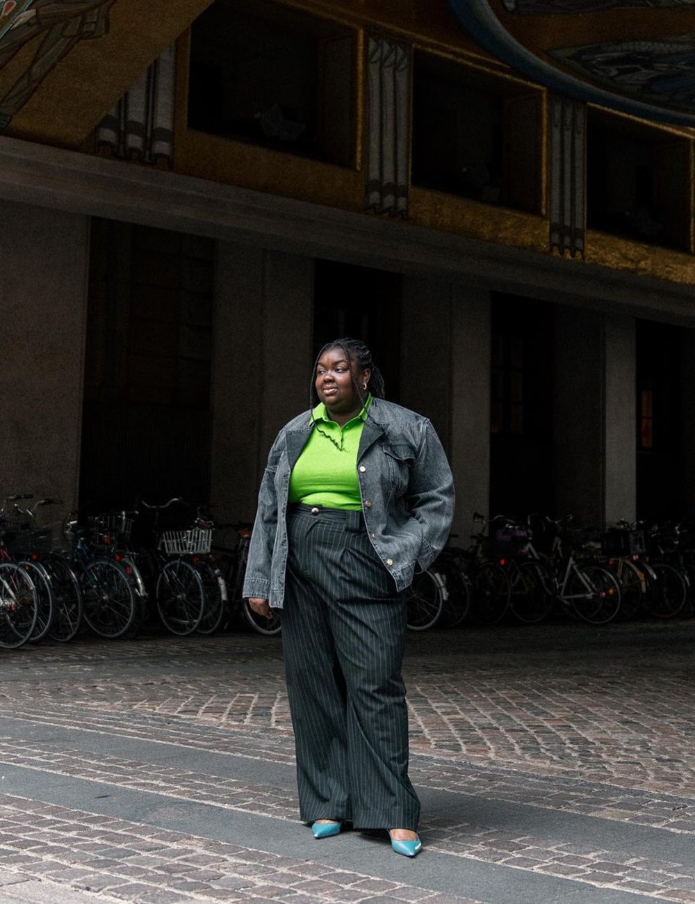 Pessoa usando jaqueta oversized de jeans cinza, blusa verde neon e calça confortável de alfaiataria com listras finas. Sapatos azuis de salto médio e cabelo trançado.