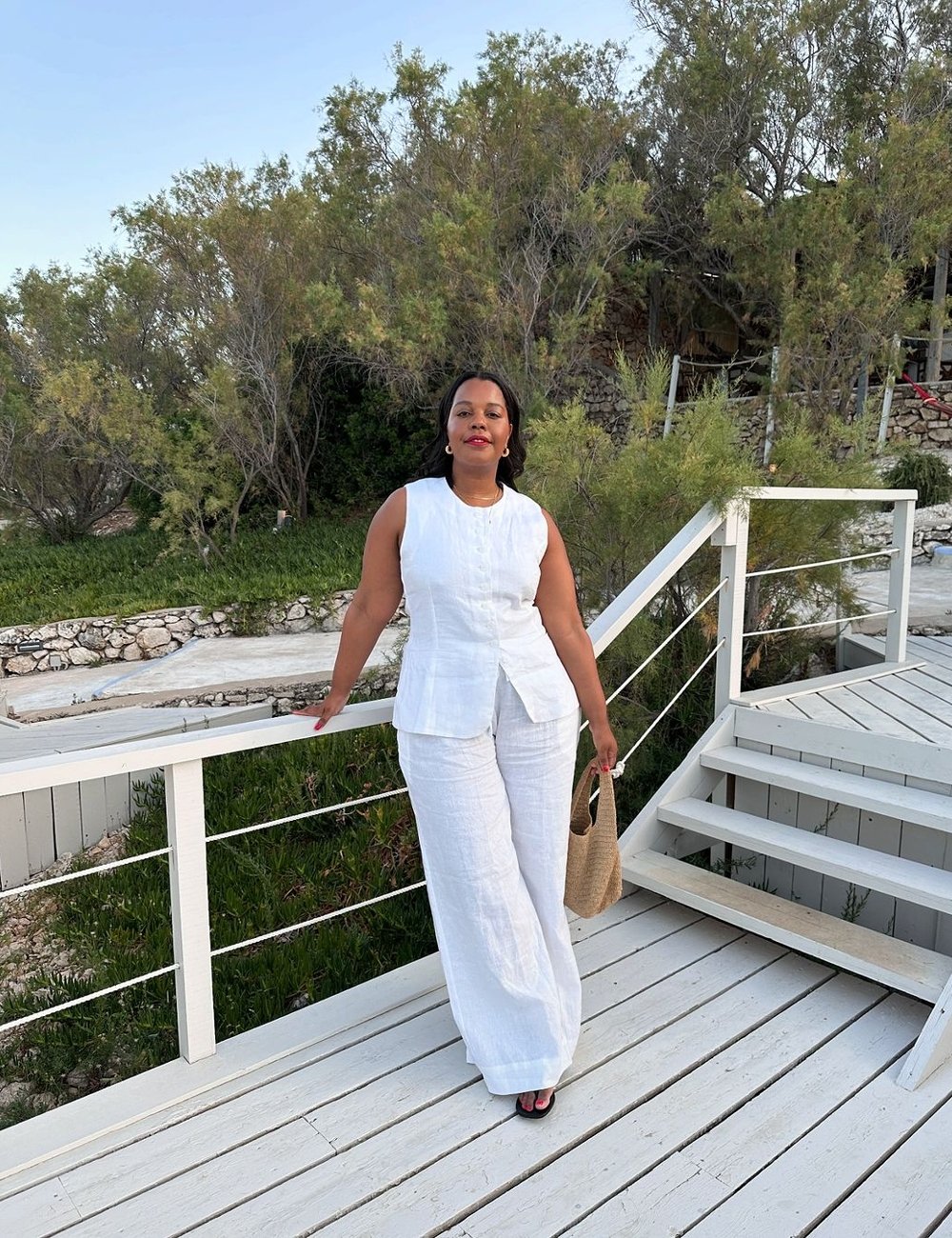 Mulher usando conjunto branco de linho, composto por calça confortável pantalona e blusa sem mangas com textura minimalista. O look é complementado por bolsa de palha e sandálias pretas, destacando uma tendência de moda de verão elegante.