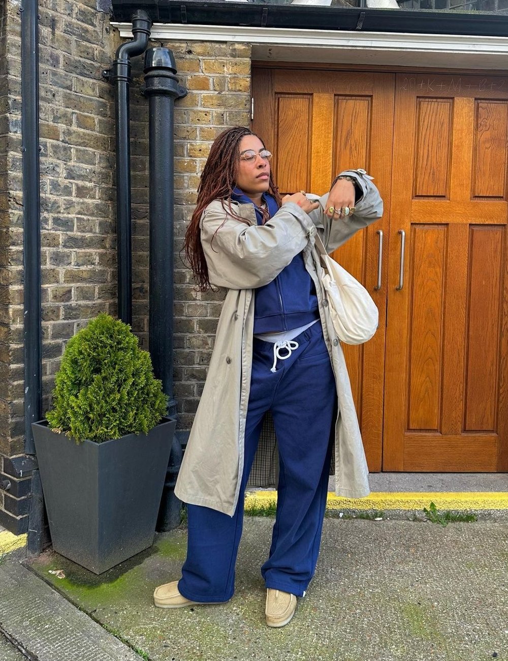 Pessoa usando look casual urbano com tendências de moda streetwear. Veste agasalho azul marinho, calça confortável, tênis bege, e um trench coat cinza longo. Acessórios incluem óculos e bolsa cruzada branca, destacando estilo despojado.