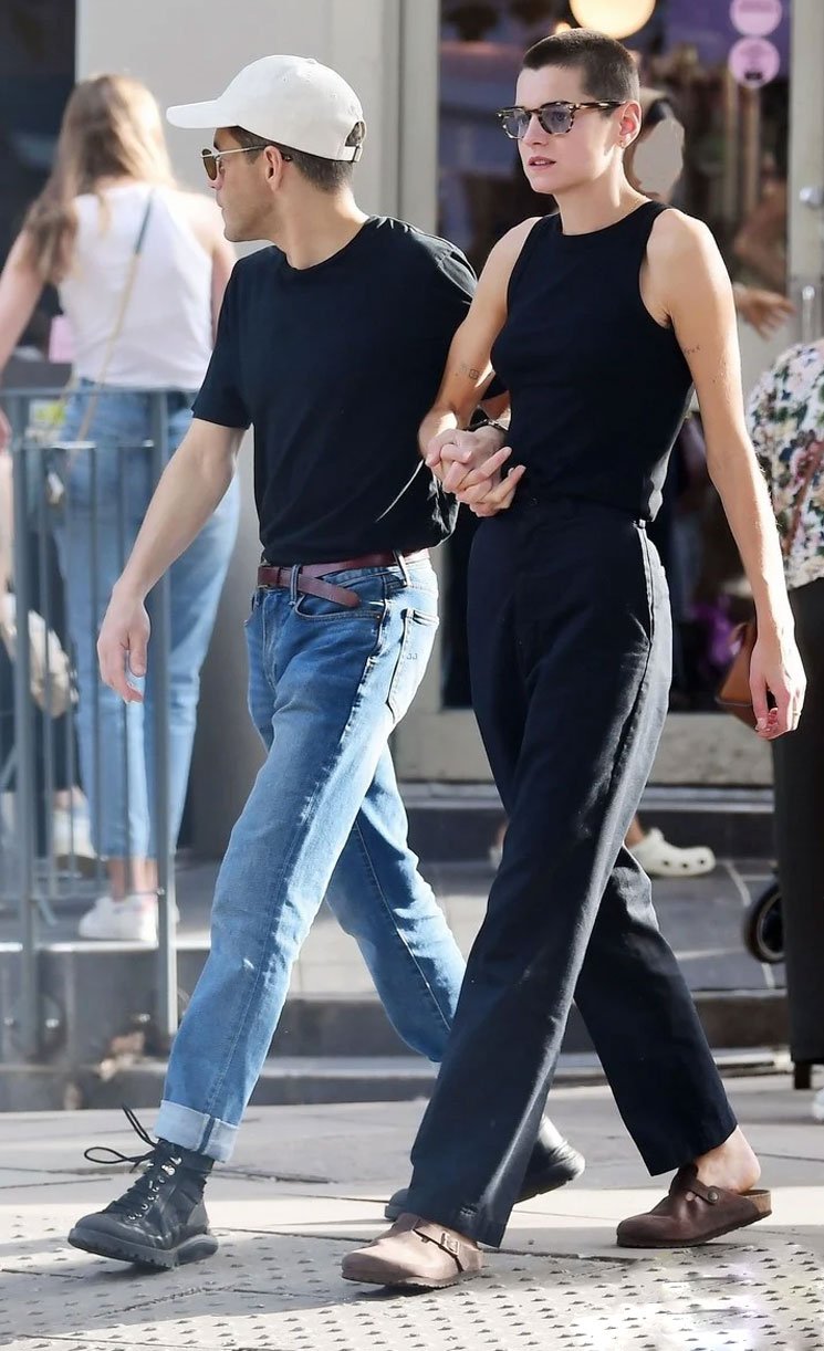 Casal caminhando na rua com looks casuais modernos. Homem veste camiseta preta, jeans com barra dobrada, tênis preto e boné branco. Mulher usa regata preta, calça larga e sapato loafer marrom. Ambos usam óculos escuros. Estilo urbano e descontraído.