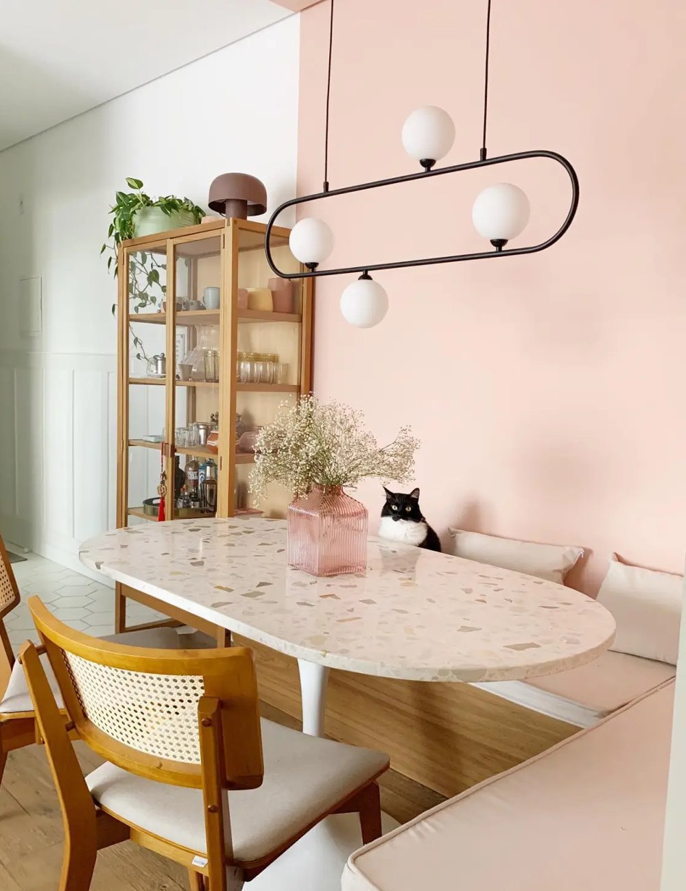 Sala de jantar moderna com canto alemão, paredes em tons pastel rosa, luminária pendente de design contemporâneo, cadeiras de madeira com estofamento claro e detalhes em palhinha. Estilo minimalista e decoração clean.