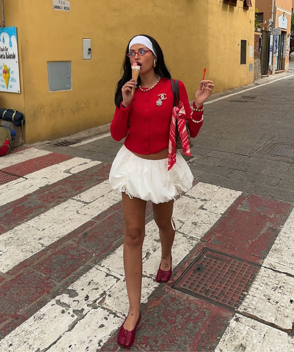 Jovem com casaco vermelho, saia branca e sapatilha vermelha. Acessoriza com bandana branca, óculos rosa e colares, destacando um look retrô e divertido. Segura bolsa vermelha com lenço e sorvete, mesclando conforto e estilo urbano.