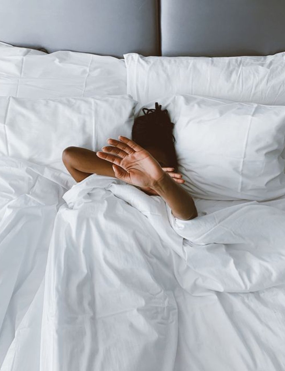 Imagem de pessoa descansando em cama com lençóis brancos de algodão. A escolha por roupas de cama neutras e confortáveis indica uma tendência minimalista e de conforto no design de interiores. Ciclos do sono.