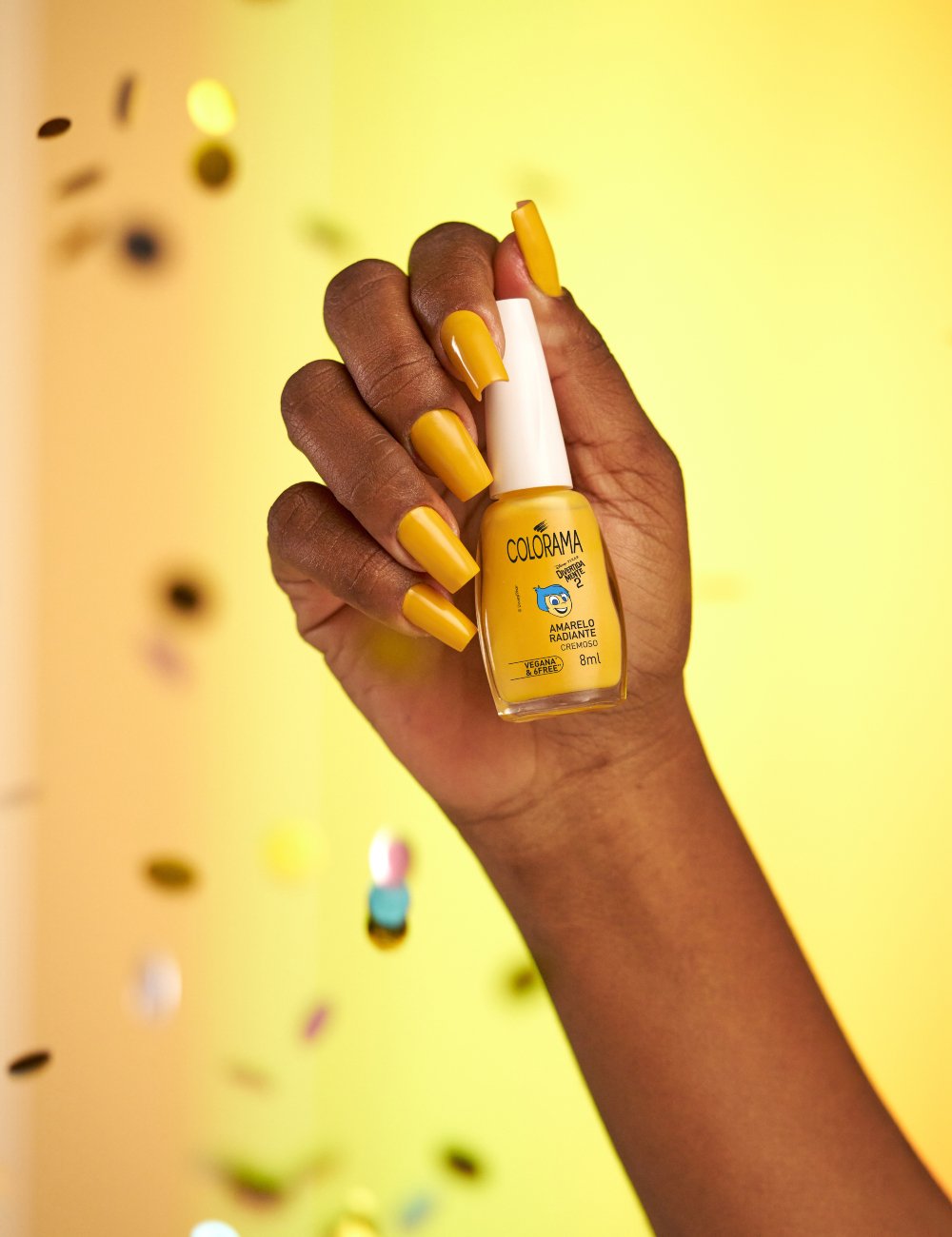 Unha com esmalte amarelo vibrante da Colorama Divertida Mente, tendência em nail art. Fundo desfocado com confetes coloridos. Ideal para looks ousados e cheios de estilo.