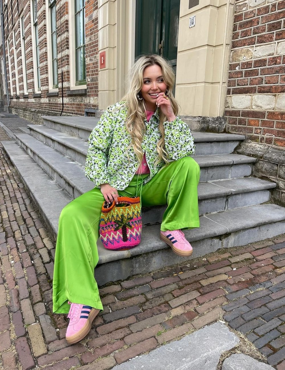 Mulher sentada em escada usando casaco estampado com flores, calça verde vibrante e tênis rosa com listras. Bolsa de crochê colorida com estampa tropical. Tendência de moda casual e criativa com combinações de cores.