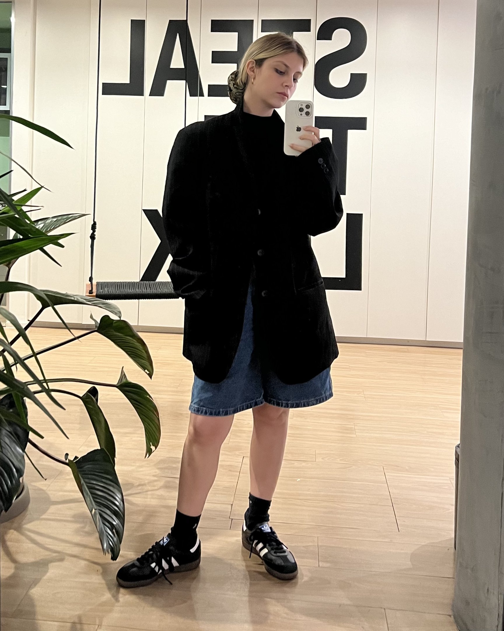 Pessoa vestindo blazer preto oversized, camiseta preta de gola alta, shorts jeans e tênis preto com branco. Look casual e moderno, combinando peças soltas e confortáveis, refletindo tendências de moda minimalista e urbana.