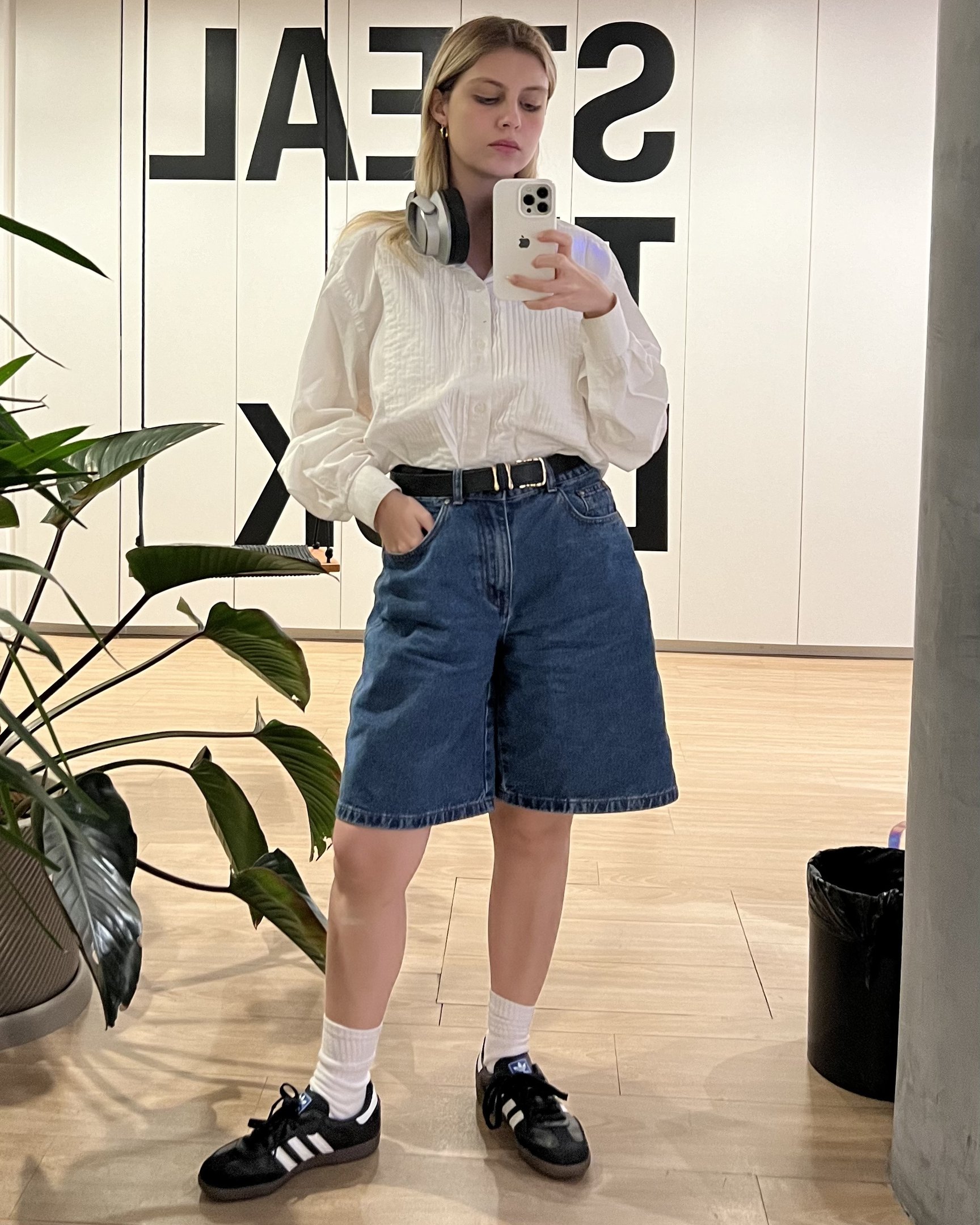 Pessoa vestindo camisa branca oversized e bermuda jeans folgada, complementando o look com um cinto preto. Calça tênis preto com meias brancas e utiliza fones de ouvido grandes no pescoço. Estilo casual e confortável, tendências de moda urbana.