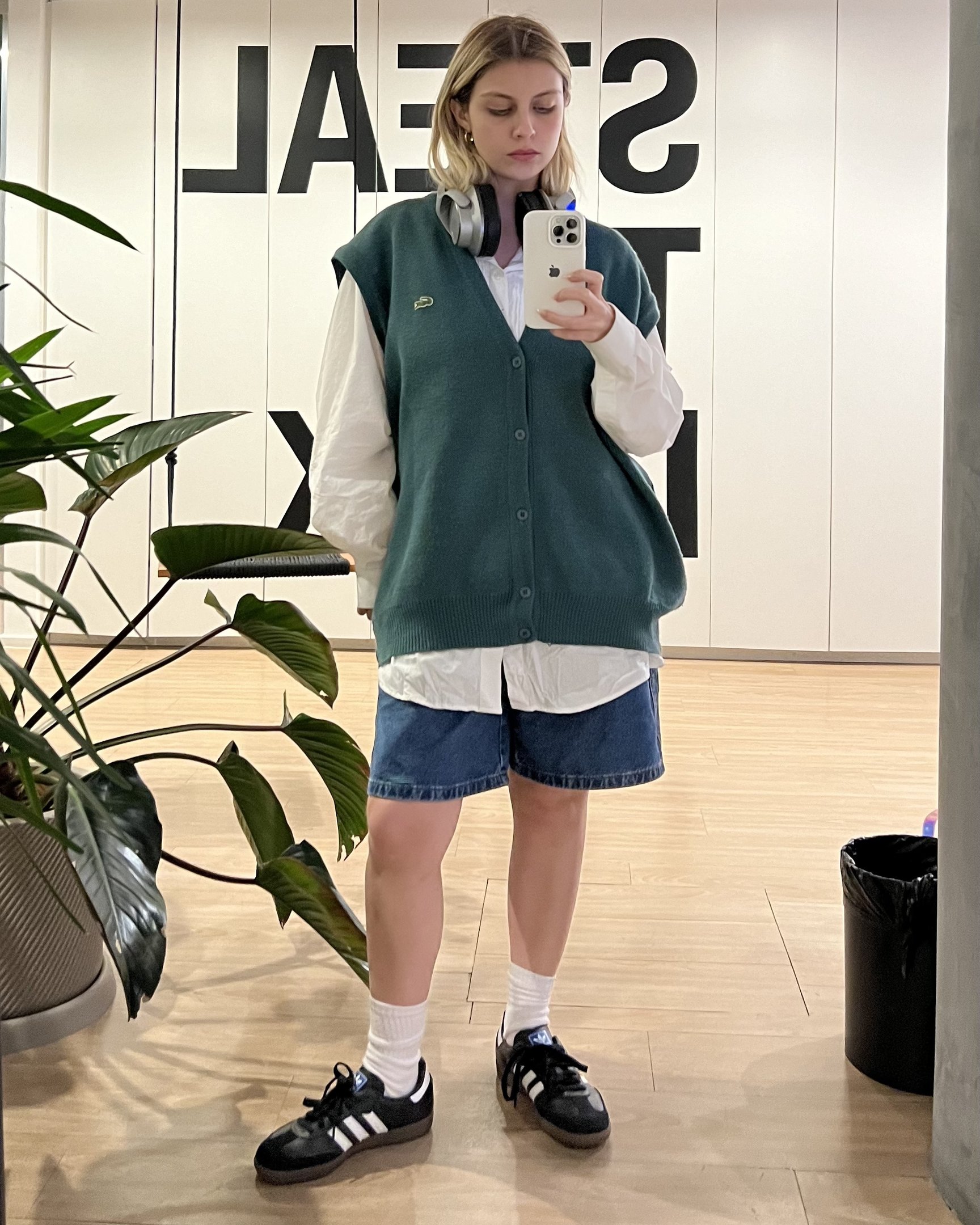 Pessoa veste camisa branca oversized com colete de tricô verde por cima, bermuda jeans e tênis Adidas samba preto. Fones de ouvido no pescoço e acessórios discretos como brincos. Estilo casual e confortável, combinando peças clássicas e esportivas.